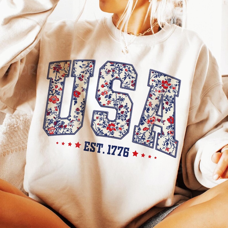May include: A white sweatshirt with a floral print of the word "USA" and the text "EST. 1776" below it.