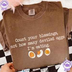 May include: A brown short-sleeve t-shirt featuring the white text "Count your blessings, not how many deviled eggs I'm eating." and a graphic of three deviled eggs with orange yolks arranged in a horizontal row below the text.