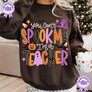 May include: A dark brown crewneck sweatshirt featuring a colorful graphic print with the text "you can't SPOOK ME I'm a TEACHER". The design includes orange, purple, and black patterned letters, a jack-o'-lantern, a ghost, a witch hat, and bats.