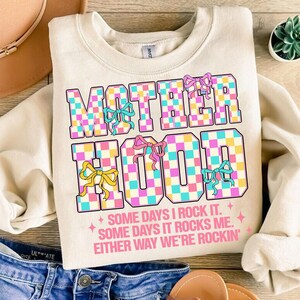 May include: A cream-coloured sweatshirt with the words "MOTHERHOOD" in pink, blue and yellow checkered letters. The letters are surrounded by pink, blue and yellow bows. The text "SOME DAYS I ROCK IT. SOME DAYS IT ROCKS ME. EITHER WAY WE'RE ROCKIN'" is printed below the checkered letters.