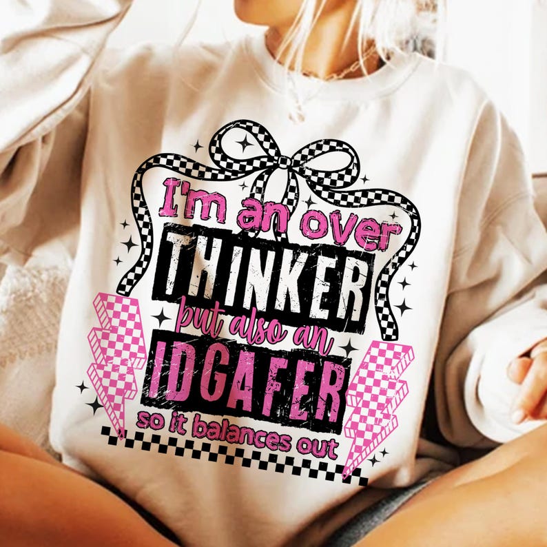 Puede incluir: Sudadera blanca con un dise&ntilde;o gr&aacute;fico. El dise&ntilde;o incluye el texto "I'm an over THINKER but also an IDGAFER so it balances out" en negro y rosa, con un lazo a cuadros y rayos.