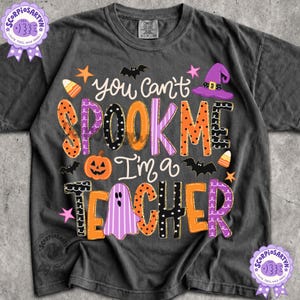 May include: A charcoal gray short-sleeve t-shirt featuring the text "you can't SPOOK ME I'm a TEACHER" in colorful, patterned lettering. The design includes orange, purple, and black graphics of a pumpkin, a ghost, bats, stars, and a witch hat.