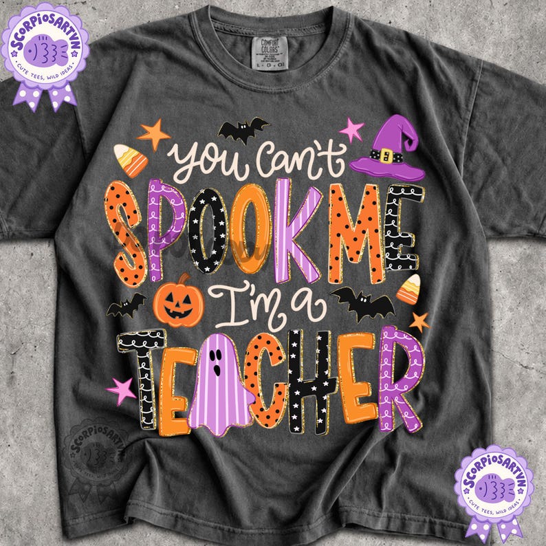 May include: A charcoal gray short-sleeve t-shirt featuring the text "you can't SPOOK ME I'm a TEACHER" in colorful, patterned lettering. The design includes orange, purple, and black graphics of a pumpkin, a ghost, bats, stars, and a witch hat.