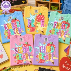 May include: Five short-sleeved crew neck t-shirts in light blue, yellow, mint green, pink, and lavender. Each shirt features a colorful graphic with a red apple, a yellow star, and text indicating school grade levels: Pre-K, Kindergarten, First, Second, and Third Grade.