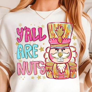 May include: White sweatshirt with the text "Y'ALL ARE NUTS" in pink and blue, with gold star accents. A colorful nutcracker graphic is on the right side, featuring pink, gold, and blue details.