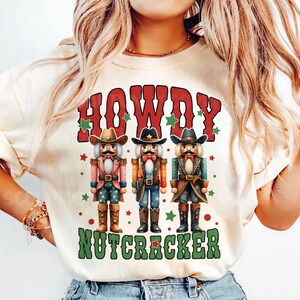 Puede incluir: Una camiseta blanca con un dise&ntilde;o rojo, verde y blanco con tres figuras de cascanueces vaquero y el texto "Howdy Nutcracker".