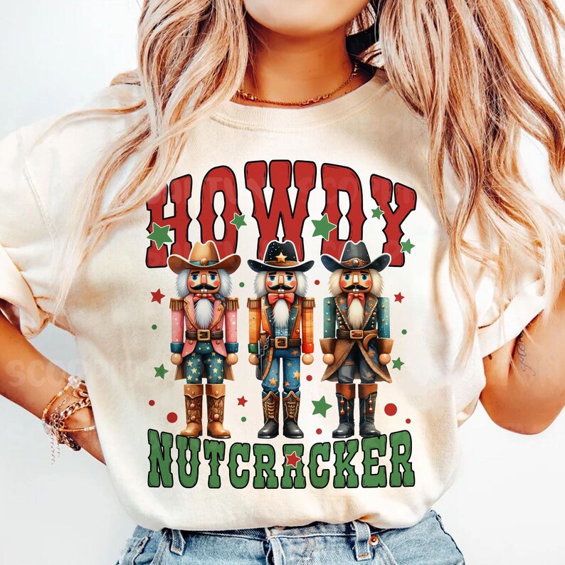 Puede incluir: Una camiseta blanca con un dise&ntilde;o rojo, verde y blanco con tres figuras de cascanueces vaquero y el texto "Howdy Nutcracker".