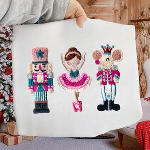 May include: A white fabric featuring embroidered Christmas characters: a nutcracker, a ballerina, and a mouse king. The figures are in pink, blue, and white, with star and crown details. The figures are approximately 6 inches tall.