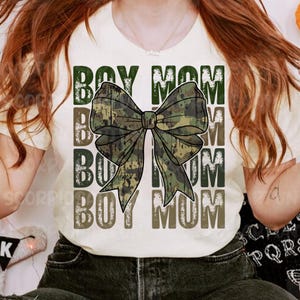 May include: White t-shirt with the text "Boy Mom" repeated four times in a distressed font. A camouflage bow is centered on the shirt.