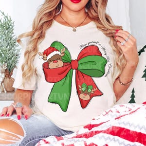 Pu&ograve; includere: Una maglietta bianca con un fiocco verde e rosso con una renna che spunta dal fiocco. Il testo "Have yourself a Merry little Christmas" &egrave; scritto sul fiocco.