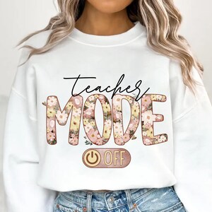 May include: White sweatshirt with the text "Teacher Mode" in floral letters with a pink and yellow color scheme. The word "Mode" is larger than the word "Teacher". Below the text is a pink and gold power button with the word "OFF" written on it.