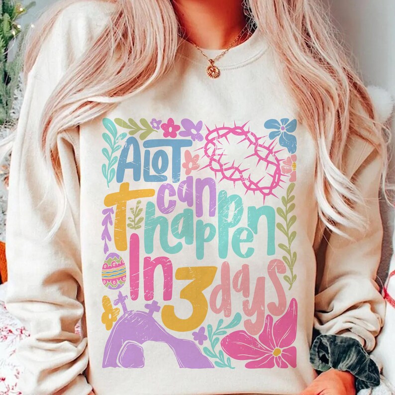 May include: A white sweatshirt with a colorful graphic design that says "A lot can happen in 3 days". The graphic includes a cross, a crown of thorns, an Easter egg, and flowers.