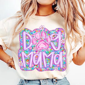 May include: A white t-shirt with a purple and pink graphic design that says "Dog Mama" with a pink paw print in the center. The graphic is surrounded by a floral pattern.