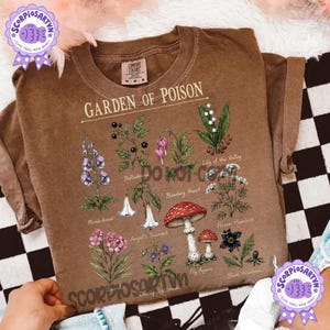 以下が含まれることがあります： Garden of Poisonというタイトルのグラフィックプリントが施された茶色の半袖Tシャツ。トリカブト、ベラドンナ、ケマンソウ、スズラン、エンゼルトランペット、ドクニンジン、ベニテングタケ、クリスマスローズなどの植物の植物画が描かれており、それぞれの名前が下に印刷されています。