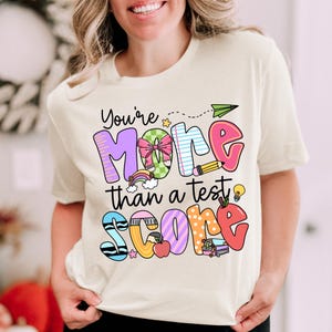 May include: A white t-shirt with a colorful graphic that says "You're More than a Test Score". The graphic includes a rainbow, a pencil, a lightbulb, a bow, and a stack of books.