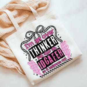 Puede incluir: Bolso tote blanco con un gr&aacute;fico en negro y rosa que dice "I'm an over thinker but also an IDGAFFER so it balances out." El bolso tiene un dise&ntilde;o de lazo y rayos.