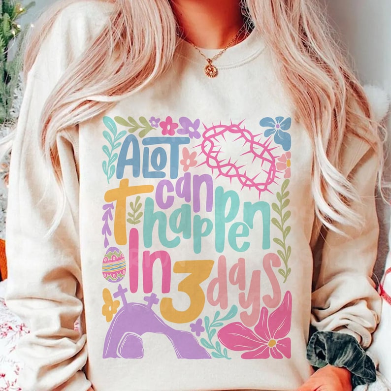 May include: A light beige sweatshirt with a colorful graphic design that says "A lot can happen in 3 days". The graphic includes flowers, a cross, a crown of thorns, and an Easter egg.