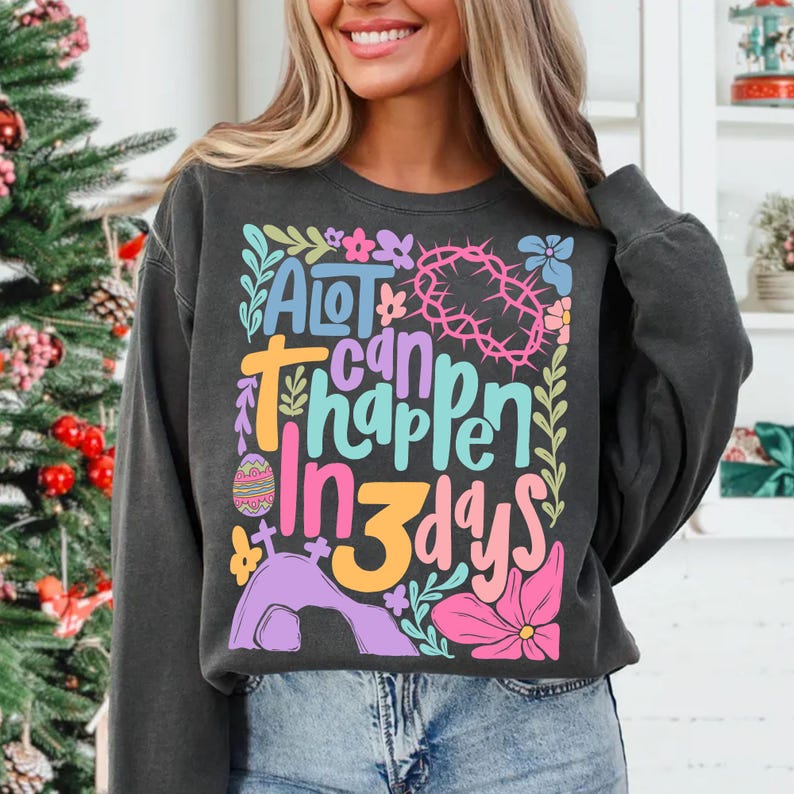 May include: A black sweatshirt with a colorful graphic design that says "A lot can happen in 3 days". The graphic includes flowers, a cross, a crown of thorns, and an Easter egg.