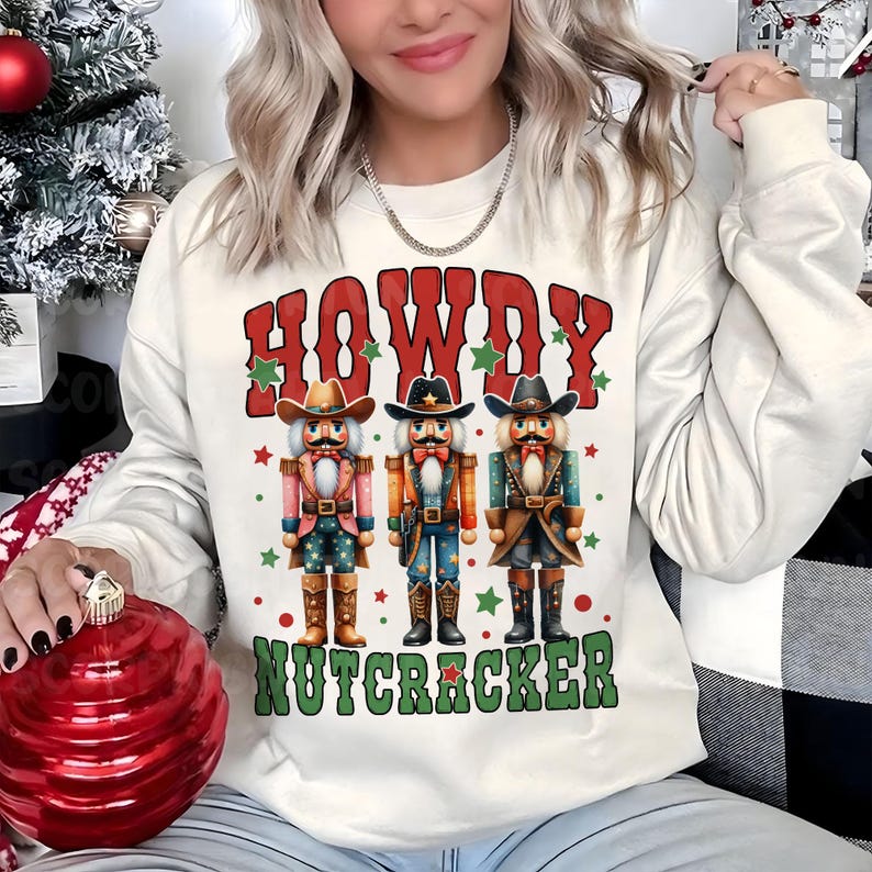 Puede incluir: Sudadera blanca con un dise&ntilde;o rojo, verde y blanco con tres figuras de cascanueces vaquero y el texto "Howdy Nutcracker".