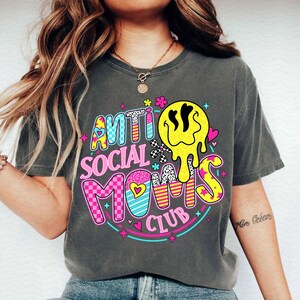 May include: Dark gray t-shirt with the text "ANTI SOCIAL MOMS CLUB" in colorful, playful lettering. The design includes a melting yellow smiley face, hearts, and a mix of patterns, creating a fun and vibrant graphic tee.