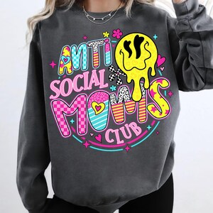 May include: Dark gray sweatshirt with the text "ANTI SOCIAL MOMS CLUB" in colorful, playful lettering. The design includes a melting yellow smiley face, hearts, and stars, creating a fun and vibrant graphic.