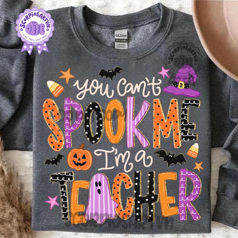 May include: A dark gray crewneck sweatshirt featuring a graphic print with the text "you can't SPOOK ME I'm a TEACHER". The design includes colorful letters with patterns, a jack-o'-lantern, a purple witch hat, bats, stars, and candy corn.