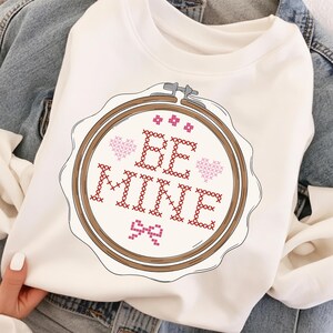 May include: A white sweatshirt featuring a cross-stitch design within a wooden embroidery hoop. The design includes the words "BE MINE" in red cross-stitch, along with pink hearts and a bow. The sweatshirt is displayed on a denim jacket.