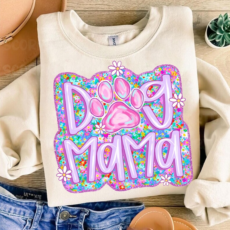 May include: A cream-colored sweatshirt with a purple and pink graphic that says "Dog Mama" surrounded by colorful flowers. The graphic includes a pink paw print.