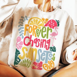 May include: A white sweatshirt with a colorful graphic design that says "Forever Chasing Sunsets". The design includes a flamingo, lemons, flowers, and leaves.