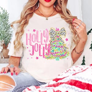 May include: White t-shirt with a pink "Holly Jolly" text and a colorful Christmas tree with a black and white polka dot bow.