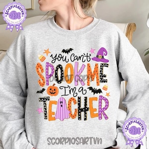 May include: A light gray crewneck sweatshirt featuring a colorful Halloween-themed graphic with the text "You can't SPOOK ME I'm a TEACHER". The design includes a pumpkin, a ghost, a witch hat, bats, stars, and candy corn in orange, purple, and black.