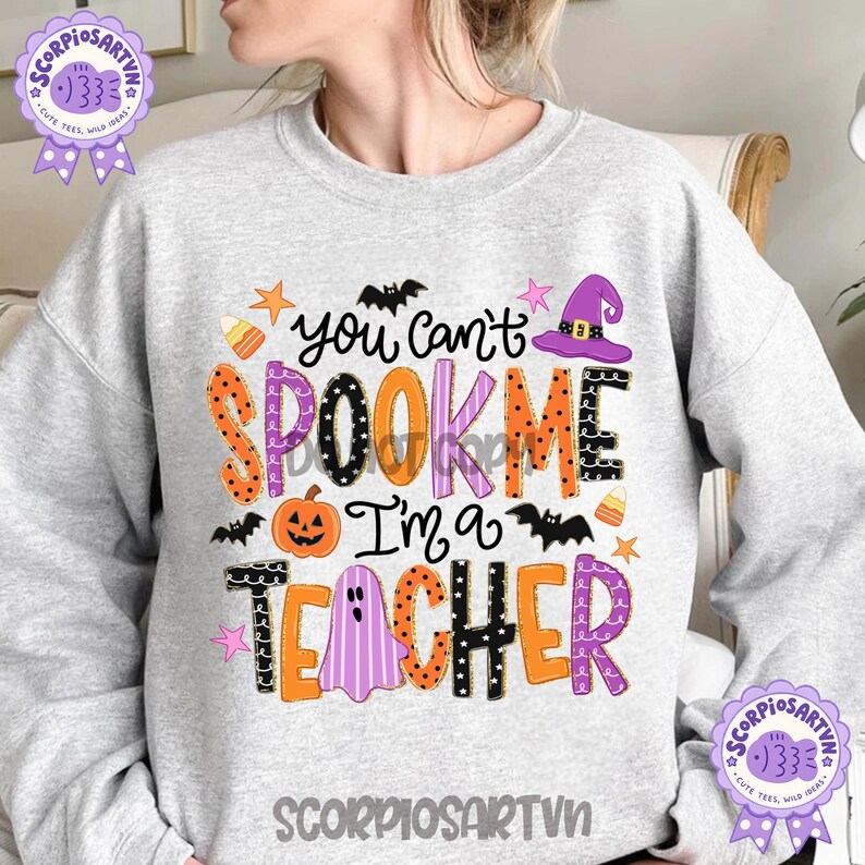 May include: A light gray crewneck sweatshirt featuring a colorful Halloween-themed graphic with the text "You can't SPOOK ME I'm a TEACHER". The design includes a pumpkin, a ghost, a witch hat, bats, stars, and candy corn in orange, purple, and black.