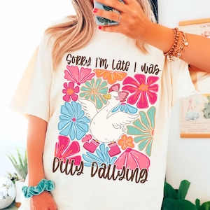 May include: A white t-shirt featuring a colourful floral design and a cartoon goose wearing pink boots. The text "Sorry I'm late I was dilly dallying" is printed on the shirt.