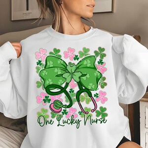 May include: White sweatshirt featuring a green bow, shamrocks, and a stethoscope design. Pink and green floral accents surround the bow and stethoscope. The text "One Lucky Nurse" is written in green script below the stethoscope.