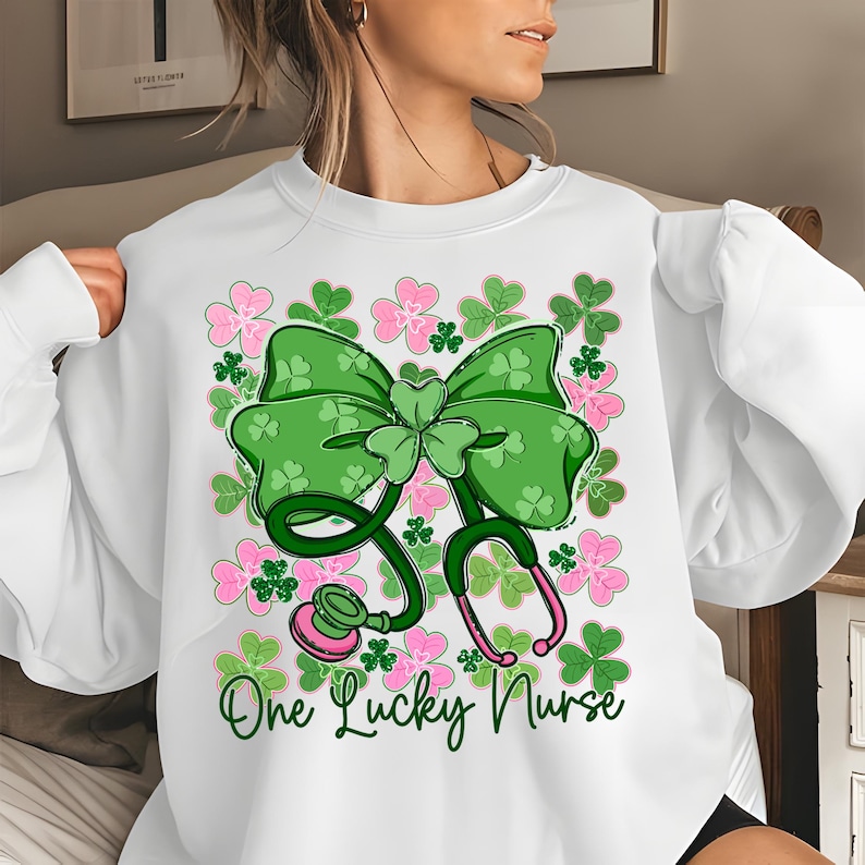 May include: White sweatshirt featuring a green bow, shamrocks, and a stethoscope design. Pink and green floral accents surround the bow and stethoscope. The text "One Lucky Nurse" is written in green script below the stethoscope.