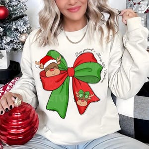 Pu&ograve; includere: Felpa bianca natalizia con un fiocco verde e rosso e due renne. Il testo sulla felpa recita "Have yourself a Merry little Christmas".