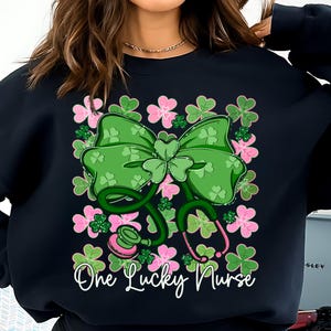 May include: Black sweatshirt featuring a green bow with shamrocks and a stethoscope design. Pink and green floral accents surround the bow and stethoscope. The text "One Lucky Nurse" is written in white script.