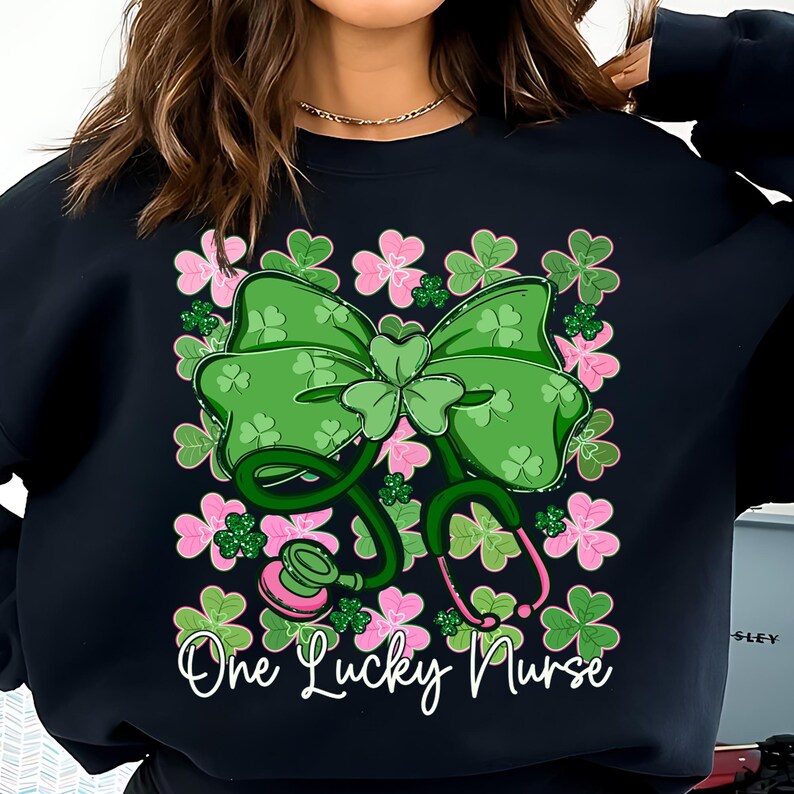 May include: Black sweatshirt featuring a green bow with shamrocks and a stethoscope design. Pink and green floral accents surround the bow and stethoscope. The text "One Lucky Nurse" is written in white script.