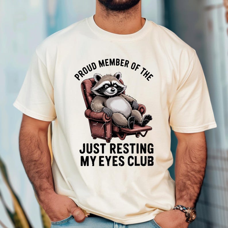 Op de afbeelding: Cr&egrave;mekleurig T-shirt met een cartoon wasbeer die ontspant in een rode fauteuil. De tekst boven de wasbeer luidt "PROUD MEMBER OF THE" en daaronder "JUST RESTING MY EYES CLUB."