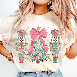 May include: A white t-shirt with a pink and green Christmas tree design featuring two nutcracker figures on either side. The tree has a large pink bow and gold glitter accents.