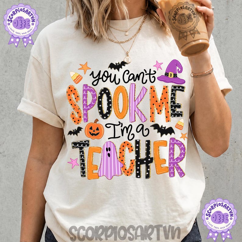 May include: An off-white t-shirt featuring the text "You can't SPOOK ME I'm a TEACHER" in colorful, patterned lettering. The design includes illustrations of a pumpkin, a ghost, bats, a witch hat, stars, and candy corn in orange, purple, and black.