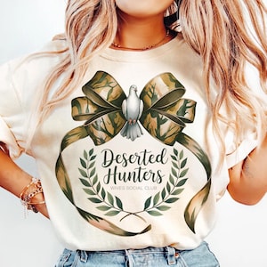 May include: A white t-shirt with a green camouflage bow and a white dove. The bow has the text "Deserted Hunters Wives Social Club" in a green wreath.