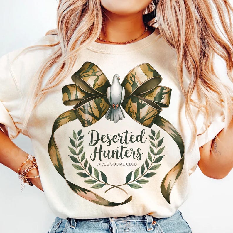 May include: A white t-shirt with a green camouflage bow and a white dove. The bow has the text "Deserted Hunters Wives Social Club" in a green wreath.
