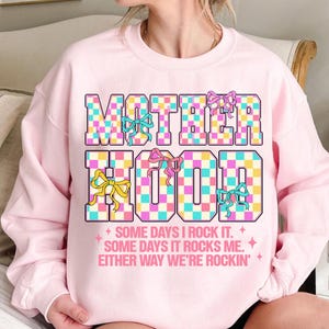May include: Pink sweatshirt with a checkered graphic that says "MOTHERHOOD" with colourful bows. The text below the graphic says "SOME DAYS I ROCK IT. SOME DAYS IT ROCKS ME. EITHER WAY WE'RE ROCKIN'."
