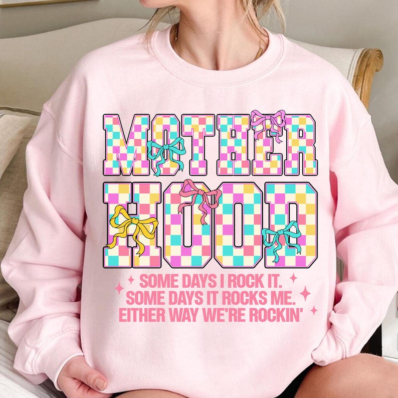 May include: Pink sweatshirt with a checkered graphic that says "MOTHERHOOD" with colourful bows. The text below the graphic says "SOME DAYS I ROCK IT. SOME DAYS IT ROCKS ME. EITHER WAY WE'RE ROCKIN'."