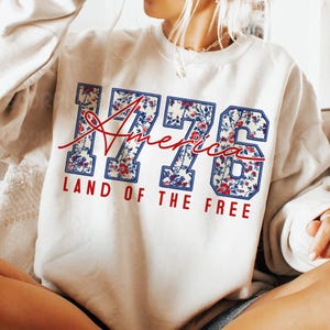 以下が含まれることがあります： 「1776 America Land of the Free」の文字が入ったクリーム色のスウェットシャツ。数字と文字は青色で、花柄と赤色のアクセントがあしらわれています。「America」の文字は筆記体です。