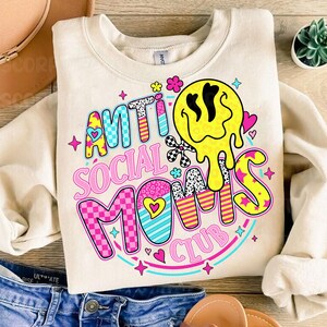 May include: Cream-colored sweatshirt with the text "ANTI SOCIAL MOMS CLUB" in colorful, playful lettering. The design includes a melting yellow smiley face, hearts, stars, and checkered patterns, creating a fun and vibrant aesthetic.
