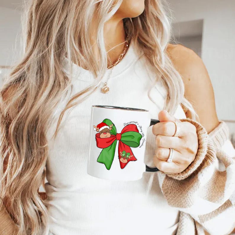 Pu&ograve; includere: Una tazza in ceramica bianca con bordo e manico neri. La tazza presenta un fiocco verde e rosso con una renna che spunta dal centro. Il testo "Have yourself a Merry Little Christmas" &egrave; stampato sulla tazza.