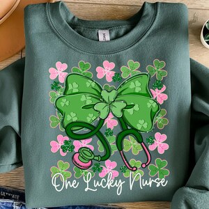 May include: A teal sweatshirt featuring a green shamrock bow and stethoscope design with pink accents. The words "One Lucky Nurse" are written in white script. The design is surrounded by pink and green shamrocks.