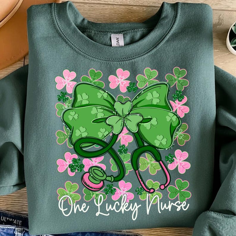 May include: A teal sweatshirt featuring a green shamrock bow and stethoscope design with pink accents. The words "One Lucky Nurse" are written in white script. The design is surrounded by pink and green shamrocks.